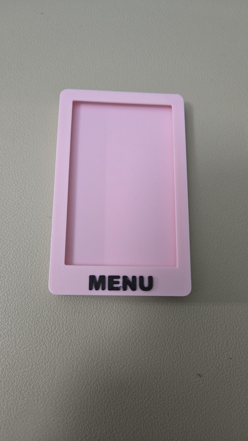 May include: A rectangular light pink plastic frame with a hollow center, featuring the word MENU in raised black capital letters at the bottom. The object has a smooth, matte finish and a simple, clean design with rounded corners on the outer edges.