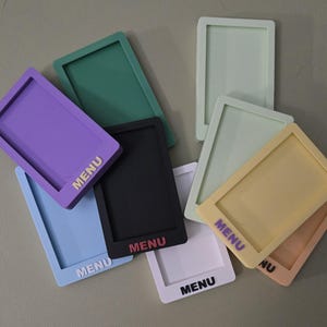 May include: A collection of rectangular silicone menu holders in various colors including purple, green, black, light blue, white, pale yellow, and peach. Each holder features a recessed center and the word MENU embossed on the bottom edge.