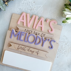 Peut inclure: Une pancarte en bois avec le texte "Avia's First Day of Melody's first day of" en lettres roses et violettes. La pancarte a un espace vide pour écrire en bas.
