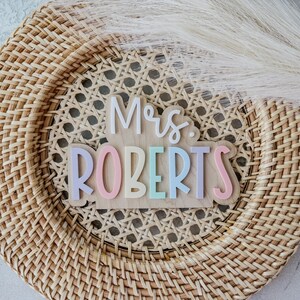 May include: A wooden sign with the text "Mrs. Roberts" in colourful letters. The letters are in different colours, including pink, yellow, blue, and green. The sign is on a woven background.