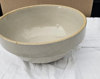 Puede incluir: Un cuenco de cerámica color crema con un diseño ligeramente inclinado. El cuenco tiene un exterior beige claro y un interior más claro. El borde tiene un aspecto más oscuro y envejecido. El cuenco está sobre una superficie blanca y texturizada.