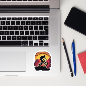 May include: A sticker with a silhouette of a person riding a mountain bike on a trail with a sunset background. The text "Be adventurous!" is written in a circle around the image.