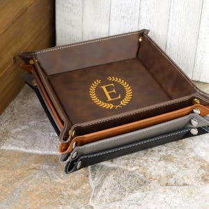 May include: A set of three leather trays in brown, tan, and gray with a monogrammed initial "E" in a gold wreath on the top tray.