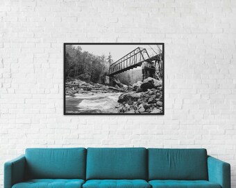 May include: Monochrome framed print of a bridge spanning a river. The metal bridge has an unusual angled structure. The fast-flowing river is surrounded by rocks and trees, creating a natural scene.