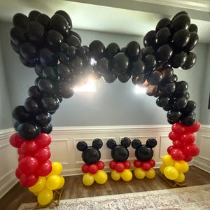 Pu&ograve; includere: Decorazione per feste con arco di palloncini e figure di palloncini. L'arco &egrave; fatto di palloncini neri, con palloncini rossi e gialli alla base. Tre figure di Mickey Mouse fatte di palloncini sono davanti.