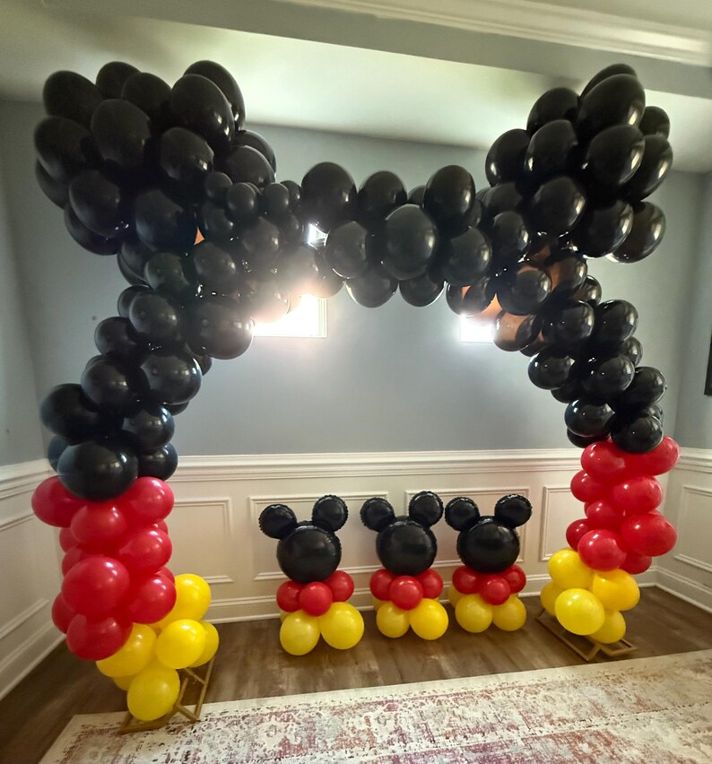 Pu&ograve; includere: Decorazione per feste con arco di palloncini e figure di palloncini. L'arco &egrave; fatto di palloncini neri, con palloncini rossi e gialli alla base. Tre figure di Mickey Mouse fatte di palloncini sono davanti.