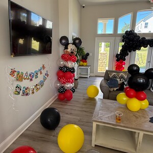 Pu&ograve; includere: Una scena di festa con decorazioni a tema Topolino. Archi e colonne di palloncini rossi, neri, gialli e bianchi creano un'atmosfera festosa. Uno striscione recita "Happy Birthday".