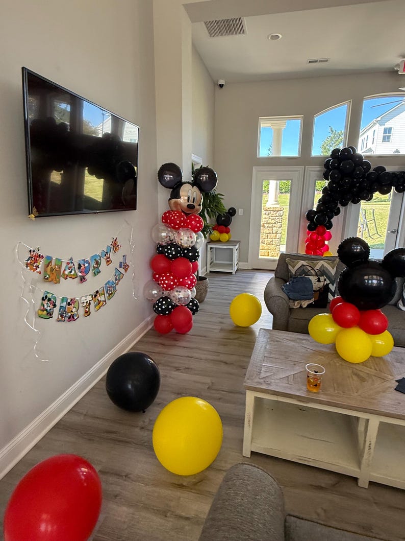 Pu&ograve; includere: Una scena di festa con decorazioni a tema Topolino. Archi e colonne di palloncini rossi, neri, gialli e bianchi creano un'atmosfera festosa. Uno striscione recita "Happy Birthday".