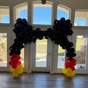 Pu&ograve; includere: Un arco di palloncini nei colori della bandiera tedesca: nero, rosso e giallo. L'arco &egrave; composto da numerosi palloncini neri che formano la parte superiore e i lati, con palloncini rossi e gialli alla base. L'arco &egrave; allestito davanti a una porta a vetri.