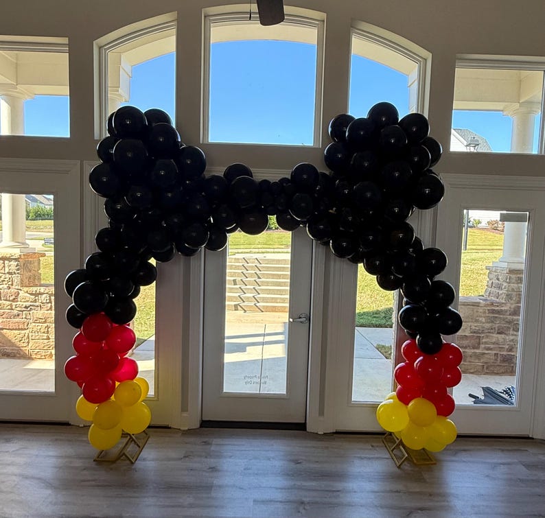 Pu&ograve; includere: Un arco di palloncini nei colori della bandiera tedesca: nero, rosso e giallo. L'arco &egrave; composto da numerosi palloncini neri che formano la parte superiore e i lati, con palloncini rossi e gialli alla base. L'arco &egrave; allestito davanti a una porta a vetri.