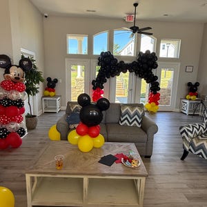 Pu&ograve; includere: Una stanza decorata con palloncini a tema Topolino. Un grande arco di palloncini neri incornicia l'ingresso. Sculture di palloncini di Topolino in rosso, nero e giallo sono collocate nella stanza. Un divano grigio e un tavolino di legno sono al centro.