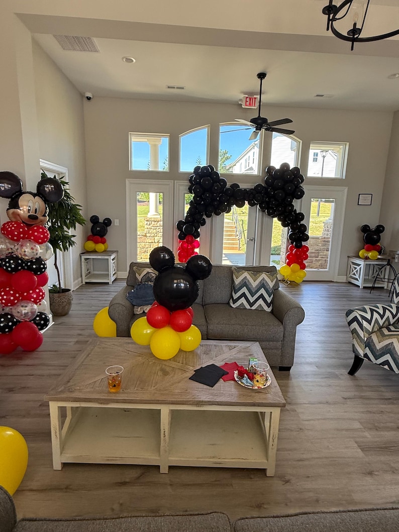 Pu&ograve; includere: Una stanza decorata con palloncini a tema Topolino. Un grande arco di palloncini neri incornicia l'ingresso. Sculture di palloncini di Topolino in rosso, nero e giallo sono collocate nella stanza. Un divano grigio e un tavolino di legno sono al centro.