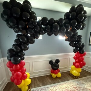 Pu&ograve; includere: Un arco di palloncini neri con accenti di palloncini rossi e gialli alla base. &Egrave; presente anche una figura di palloncino di Topolino. I palloncini sono disposti su supporti di legno, creando una decorazione festosa per feste.