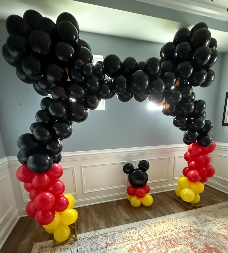 Pu&ograve; includere: Un arco di palloncini neri con accenti di palloncini rossi e gialli alla base. &Egrave; presente anche una figura di palloncino di Topolino. I palloncini sono disposti su supporti di legno, creando una decorazione festosa per feste.