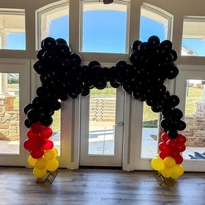 Pu&ograve; includere: Un arco di palloncini neri a forma di fiocco, con palloncini rossi e gialli alla base. L'arco &egrave; posizionato in una stanza con grandi finestre, creando un ingresso festoso e decorativo per un evento.