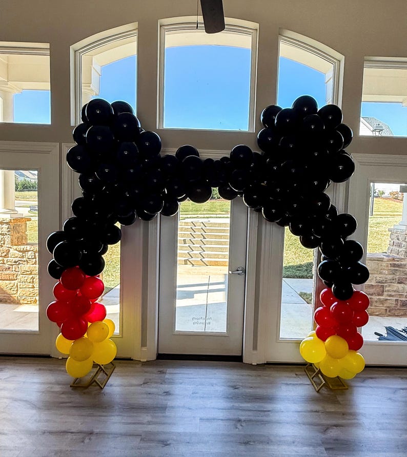 Pu&ograve; includere: Un arco di palloncini neri a forma di fiocco, con palloncini rossi e gialli alla base. L'arco &egrave; posizionato in una stanza con grandi finestre, creando un ingresso festoso e decorativo per un evento.