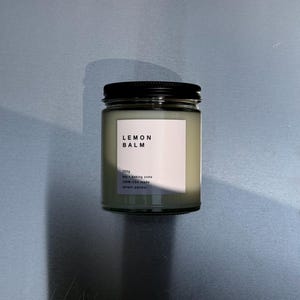 May include: A glass jar with a black lid, labelled "LEMON BALM." The label also indicates the contents are 250g, made with soy and baking soda, and 100% USA made. The candle has a pale green hue.
