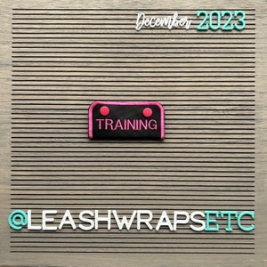 May include: Black and pink fabric patch with the word "TRAINING" in white lettering. The patch has two pink circles on the top. The patch is on a wood background with the text "December 2023" and "@LEASHWRAPSETC" in white and turquoise letters.