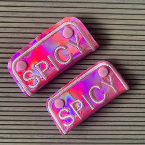 May include: Two rectangular, iridescent pouches with the word "SPICY" embroidered in white. Each pouch has a pink border and two pink circular fasteners. The pouches are on a gray, ridged surface.
