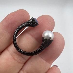 May include: Black ring with a purple gemstone and a white pearl. The ring is designed with intricate details and has a unique shape.