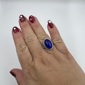 May include: A silver ring with a large oval blue gemstone. The ring is on a person's finger. The person's fingernails are painted a dark red color with white polka dots.