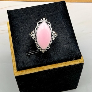 May include: A silver ring featuring a marquise-cut, smooth, light pink stone set in an ornate, filigree-style metal frame. The setting has intricate, decorative metalwork surrounding the central pink gemstone, creating a detailed, symmetrical design.