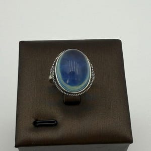 May include: A silver ring with an oval, blue gemstone. The gemstone has a translucent appearance with a lighter blue center. The ring is set on a brown display box against a white background. The ring's setting has a detailed, rope-like design.