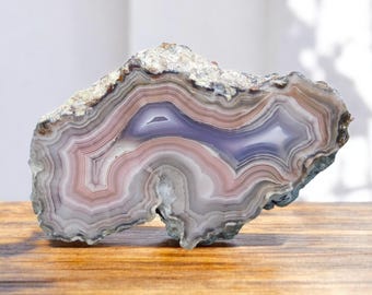 Pode incluir: Uma fatia de ágata polida com um padrão de espiral de bandas rosa, brancas e roxas. A ágata é uma pedra natural com uma superfície lisa e polida.