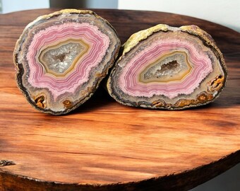 May include: Two halves of a polished agate geode, displaying concentric bands of pink, white, and brown. The geodes are presented on a wooden surface, revealing a natural, layered interior with a central cavity.
