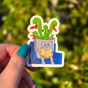 May include: A sticker featuring a cartoon plant in a gray pot, wearing glasses and reading a yellow book while seated on a blue couch. The plant has green leaves with red accents. The background is blurred green.