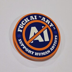 May include: A stack of round stickers with a blue and orange design. The stickers have the text "FUCK AI 'ART' SUPPORT HUMAN ARTISTS" in white. The center of the sticker has a white "AI" symbol crossed out with an orange line.