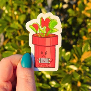 May include: A sticker featuring a cartoon Venus flytrap in a red pot with an angry face. The plant has red and green leaves. The pot is holding a small, rectangular object. The background is blurred green foliage.