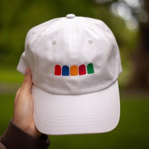 Puede incluir: Gorra de béisbol blanca con visera curva. La gorra presenta un diseño bordado de cinco formas arqueadas de colores en rojo, azul, naranja, rojo y verde. La gorra está sujeta por una persona.