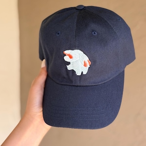 Könnte beinhalten: Marineblaue Baseballkappe mit einem gestickten Design einer weißen und roten Kreatur auf der Vorderseite. Die Kappe hat einen gebogenen Schirm und einen Stoffriemenverschluss. Ein stilvolles Accessoire.