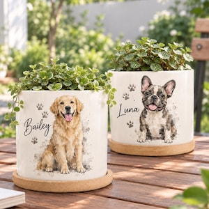 Puede incluir: Dos macetas cilíndricas de cerámica blanca con platos de madera, cada una con una planta verde. Una maceta presenta una ilustración de un golden retriever con el nombre Bailey, y la otra muestra un bulldog francés con el nombre Luna y huellas.