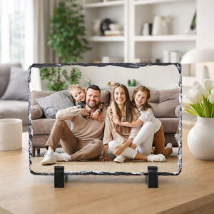 Op de afbeelding: Een rechthoekige fotoafdruk op leisteen met een familieportret. De afbeelding toont een lachende familie van vier, met een man, een vrouw en twee kinderen. De leisteen wordt ondersteund door twee zwarte standaards en staat op een houten tafel.