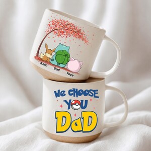 May include: Two white ceramic mugs stacked on top of each other. The top mug features a cartoon scene with a tree and three characters. The bottom mug has the text "We Choose You Dad" with a Pokeball graphic.
