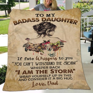 May include: A brown and tan fleece blanket with a motivational message for a daughter. The blanket features a drawing of a woman with butterflies on her shoulders and the text "To my badass daughter. If fate whispers to you "You can't withstand the storm" whisper back "I am the storm." Wrap yourself up in this and consider it a big hug. Love, Dad."