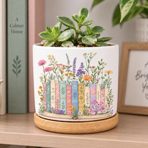 Puede incluir: Una maceta cilíndrica de cerámica blanca con una ilustración colorida de una fila de libros de cuyas páginas brotan flores silvestres. La maceta se asienta sobre un plato circular de madera clara y contiene una pequeña planta suculenta verde.