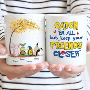 May include: Two white ceramic mugs. One mug features cartoon characters under a tree with yellow leaves and names. The other mug has the text "Catch 'Em All but keep your FRIENDS CLOSER" in yellow and blue.