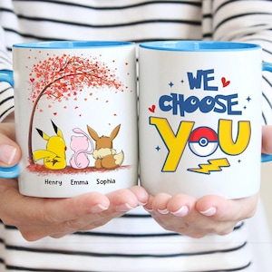 May include: Two white ceramic mugs with blue handles and rims. One mug features Pikachu, Mew, and Eevee under a tree with the names Henry, Emma, and Sophia. The other mug says "We Choose You" with a Pok&eacute; Ball design.