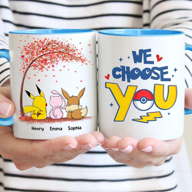 May include: Two white ceramic mugs with blue handles and rims. One mug features Pikachu, Mew, and Eevee under a tree with the names Henry, Emma, and Sophia. The other mug says "We Choose You" with a Pok&eacute; Ball design.