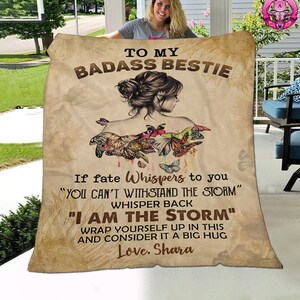 May include: A tan and brown throw blanket with a quote about being strong and a woman with butterfly wings. The quote reads "To my badass bestie, If fate whispers to you "You can't withstand the storm" whisper back "I am the storm" Wrap yourself up in this and consider it a big hug. Love, Shara"