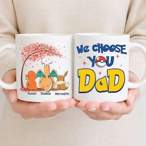 May include: Two white ceramic mugs. One mug features a tree with red leaves and three cartoon characters with the names "Nolan", "Daddy", and "Meredith". The other mug says "We Choose You Dad" with a Pokeball.