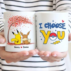 Op de afbeelding: Twee witte keramische mokken. Eén mok toont Pikachu en Eevee onder een boom met rode bladeren, met de namen "Henry" en "Emma". De andere mok zegt "I Choose You" met een Pokeball-ontwerp.