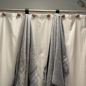 May include: Two gray towels hanging on a shower curtain rod with white shower curtains. The towels are hanging from decorative hooks with rope accents.