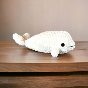 May include: White crocheted stuffed beluga whale toy with black eyes. The toy is lying on a wooden surface.