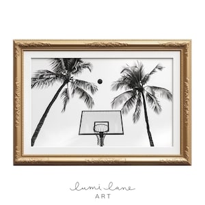 May include: Black and white photograph of a basketball hoop with a basketball in the air, framed by two palm trees. The image is framed in a gold frame.