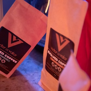 May include: Two brown paper bags with black labels that read "Victor in Valor Evolved Coffee" and "High Flying Blend".