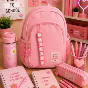 Op de afbeelding: Een roze rugzak met een gepersonaliseerd naamlabel met de tekst MARLEEY, een roze waterfles met een smiley, twee spiraalnotitieblokken met tekst, een roze etui en een roze pennenhouder van gaas met diverse roze pennen en stiften.